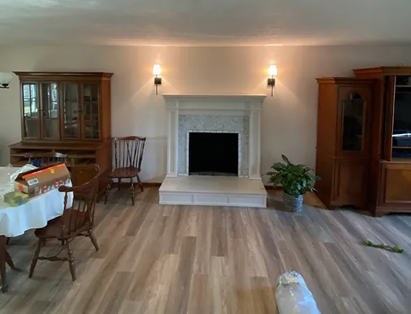 A cozy living room with a white fireplace centered on a light wood floor. Two wall sconces illuminate the space. Surrounding are chairs, a dining table, and wooden cabinets, creating a warm, inviting atmosphere.